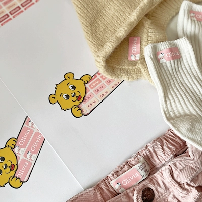 Folha de etiquetas cor-de-rosa com um autocolante de urso, ao lado de uma camisola amarela, meias brancas e calções cor-de-rosa