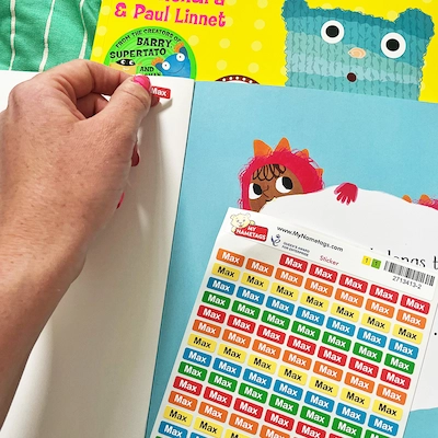 Mão a descolar um mini autocolante de uma folha, ao lado de um livro infantil ilustrado