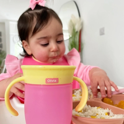 Bebé a beber de um copo cor-de-rosa com um mini autocolante com o nome Olivia