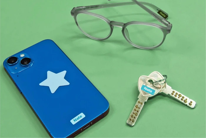 Telemóvel azul, óculos e chaves sobre fundo verde, cada um com um mini autocolante com o nome Nate