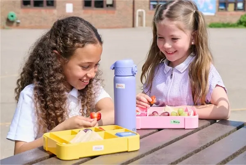 Duas meninas de uniforme escolar a lanchar ao ar livre, com uma garrafa roxa e lancheiras etiquetadas