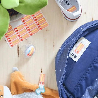 Folhas de etiquetas cor de laranja sobre uma mesa, com uma mochila, uma chupeta e peluches